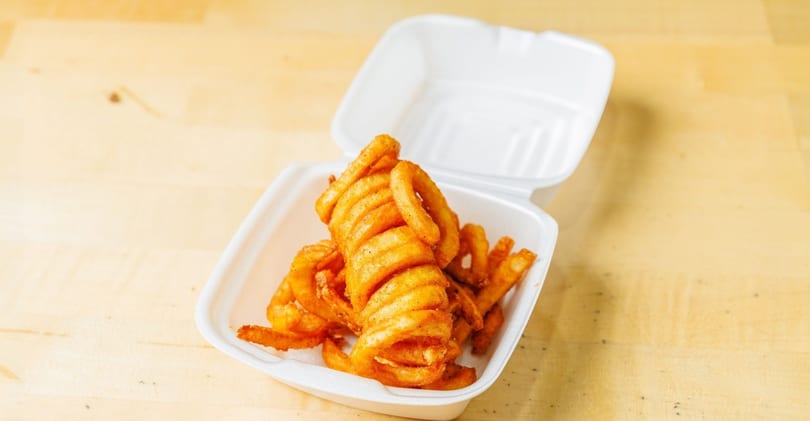 Seasoned curly fries in a white styrofoam takeout container on a light wooden surface.