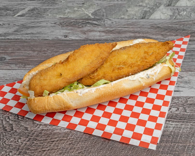 A fried fish po'boy sandwich with lettuce, tomato, and tartar sauce on a red and white checkered paper.
