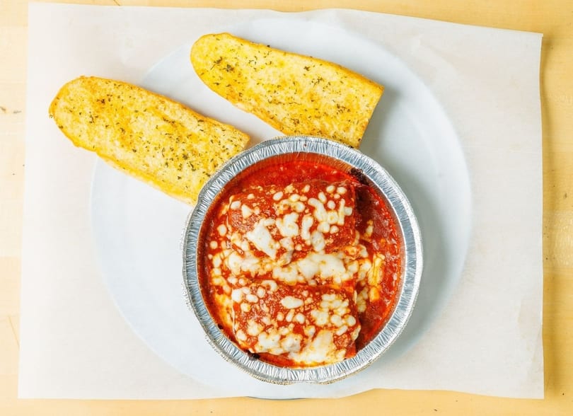 Baked ziti with melted cheese and tomato sauce in a foil container, served with two garlic breadsticks.