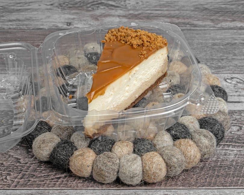 A slice of cheesecake with caramel topping and cookie crumbles in a plastic container.