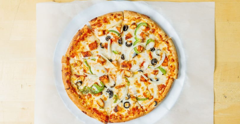 A whole pizza with chicken, green peppers, onions, and black olives on a white paper.