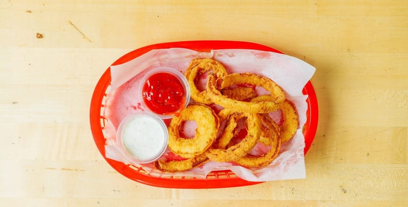 Golden-brown onion rings in a red basket with ketchup and ranch dipping sauces on a wooden table.