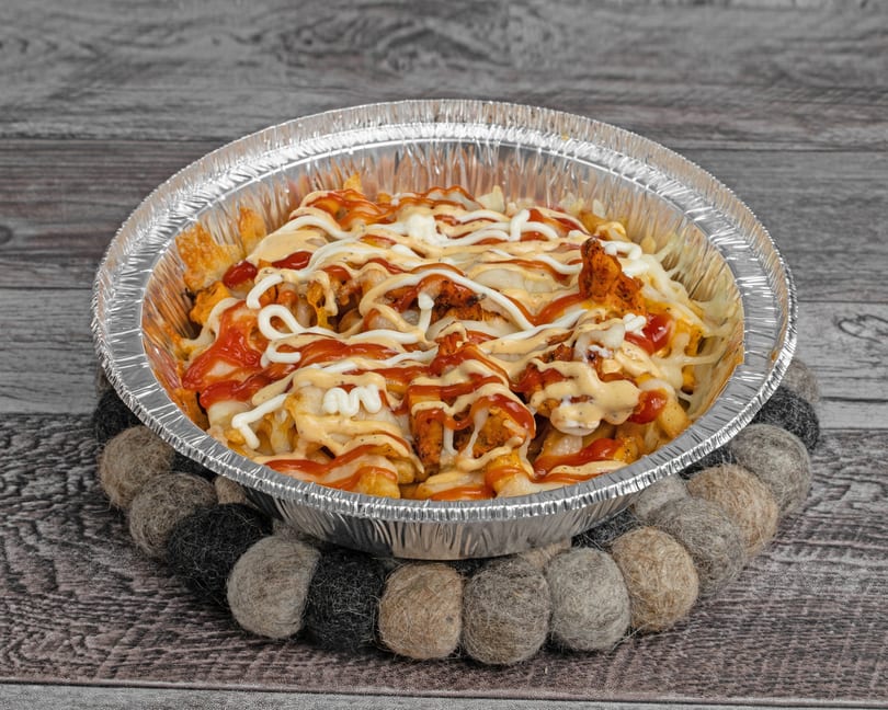 Fries topped with chicken, cheese, ketchup, and white and orange sauces in a foil container.