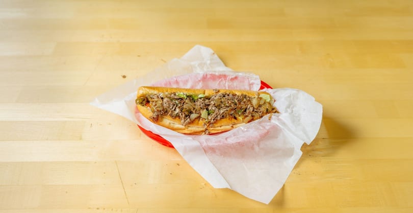 A large cheesesteak sandwich with steak, onions, and peppers in a red basket on a wooden table.