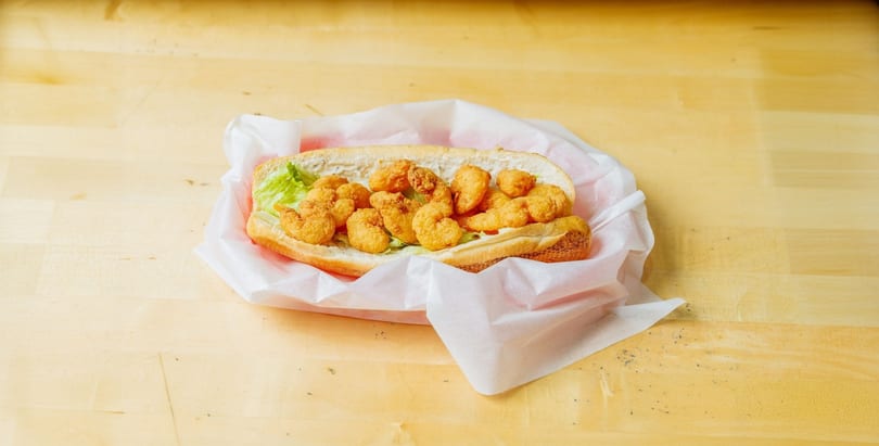 A shrimp po'boy sandwich with lettuce and sauce, wrapped in white paper on a wooden table.