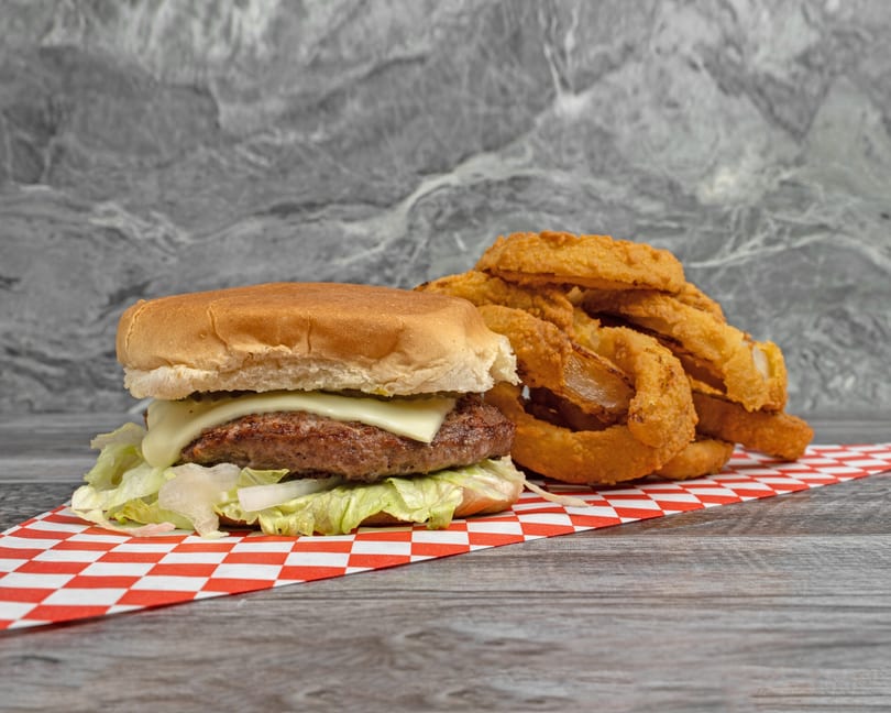 Cheeseburger with lettuce and onions, alongside a pile of golden onion rings on a checkered paper.