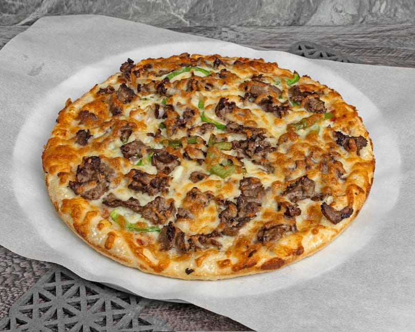 A whole round pizza with melted cheese, steak, and green peppers on a white paper liner.